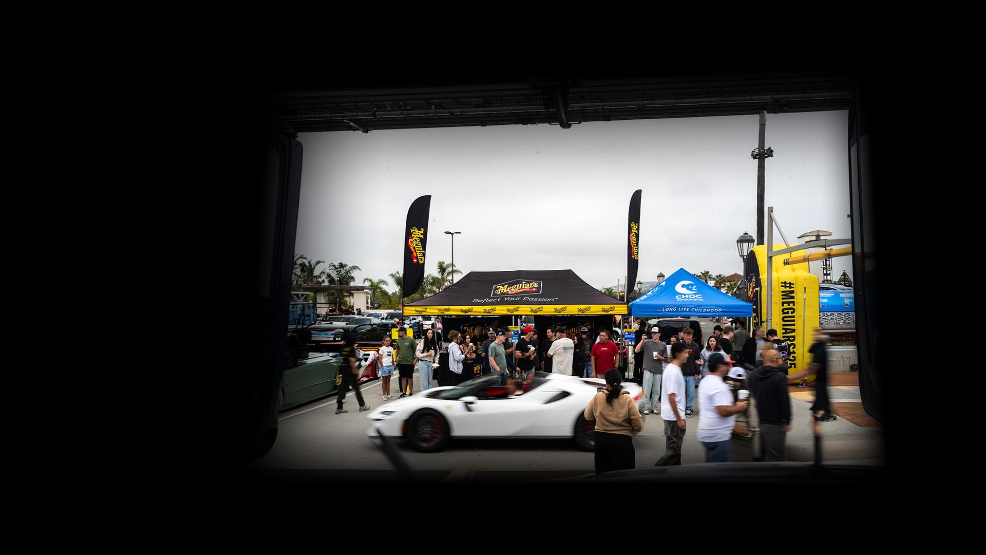 People at a car show with a white sports car in front of the Meguiar's booth