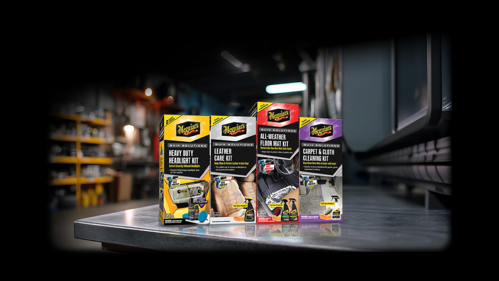 Four Meguiar’s car care product kits displayed on a metal surface in a workshop setting. Kits include Heavy Duty Headlight Kit, Leather Care Kit, All-Weather Floor Mat Kit, and Carpet & Cloth Cleaning Kit.