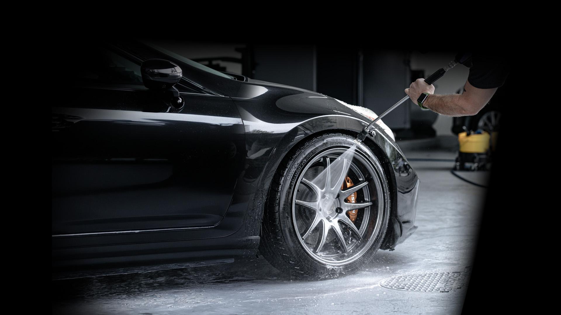 A close-up of a black car's front wheel being cleaned with a high-pressure water spray. The image highlights water droplets and foam on the tire and rim.