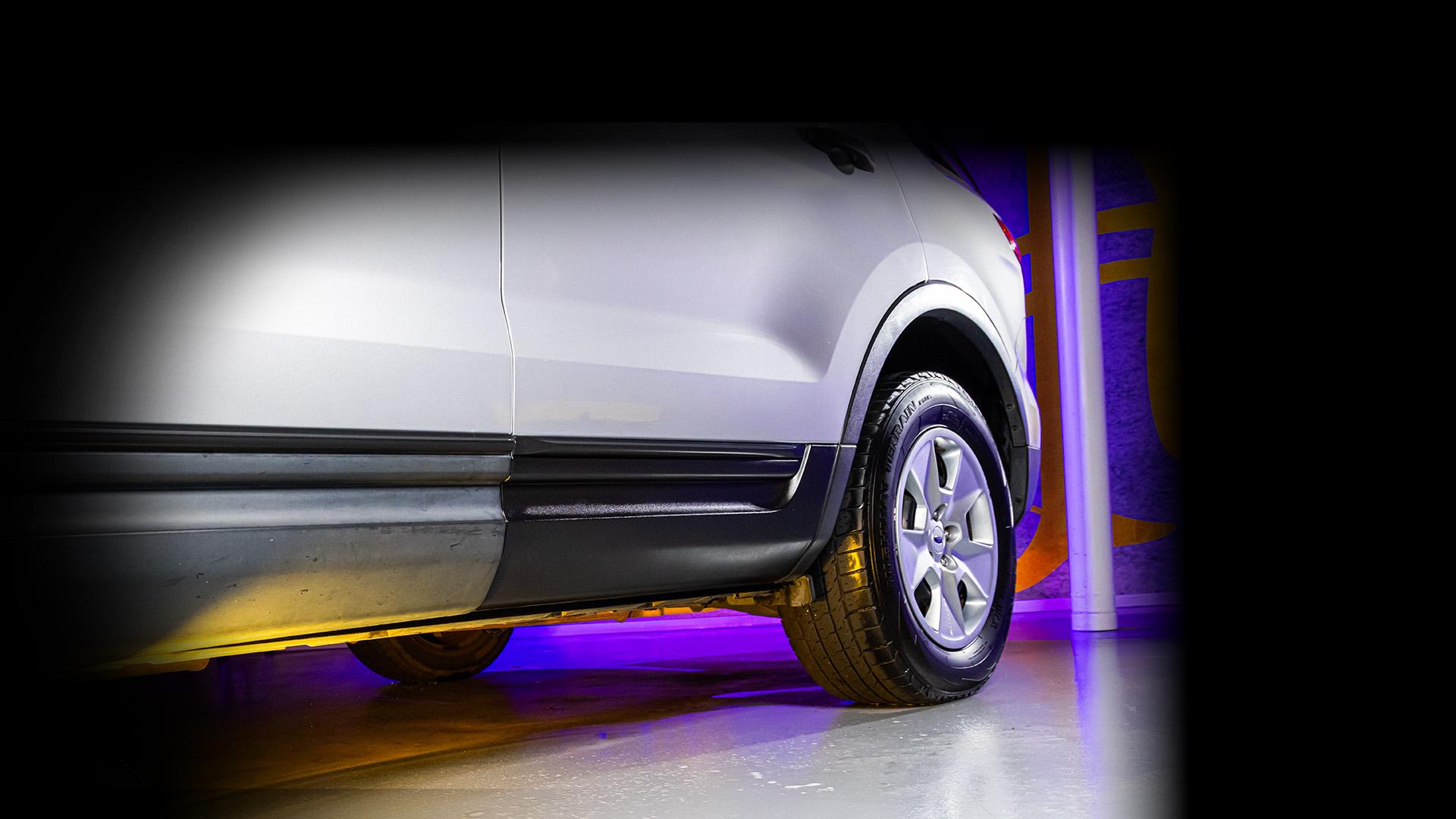 A close-up side view of a white vehicle's rear tire and lower body panel, with a glossy finish reflecting purple and yellow lighting. The image highlights the tire tread, wheel design, and clean undercarriage, emphasizing vehicle condition and detailing.