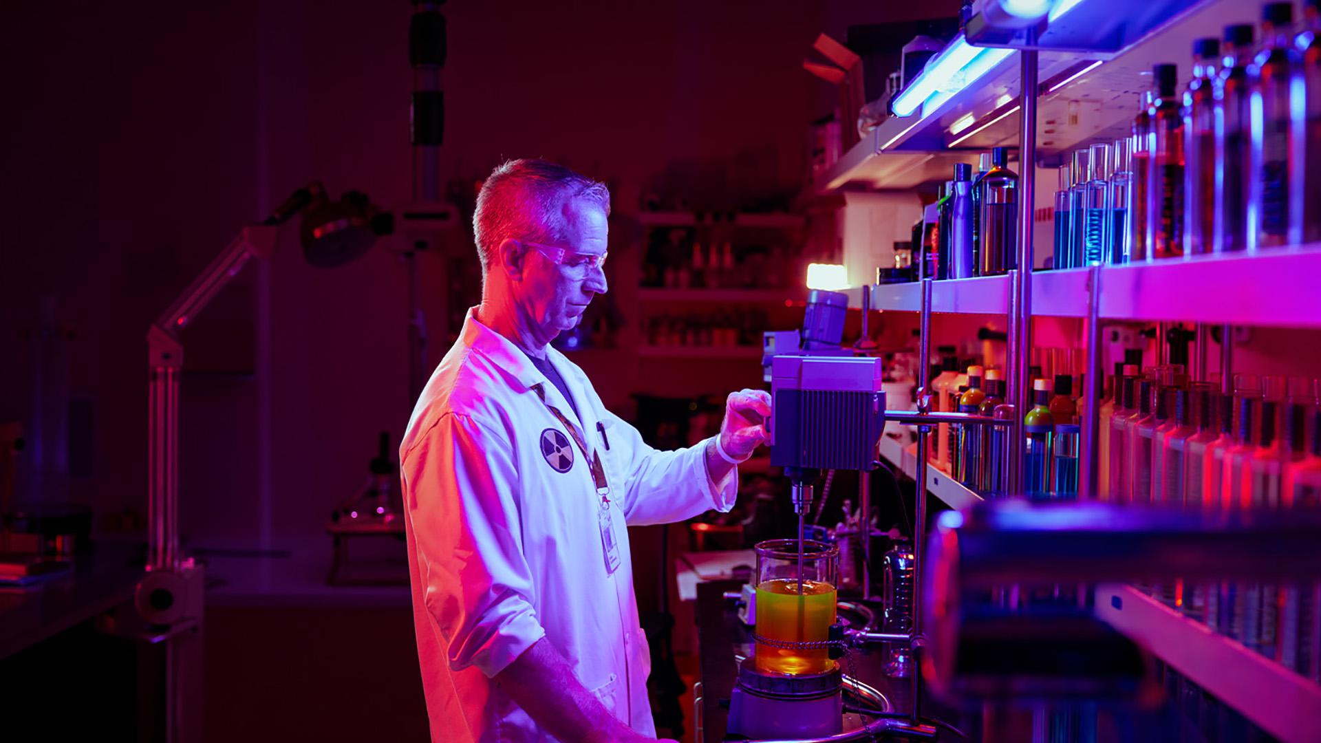 Person in white lab coat using lab equipment with shelves of chemicals in front of him