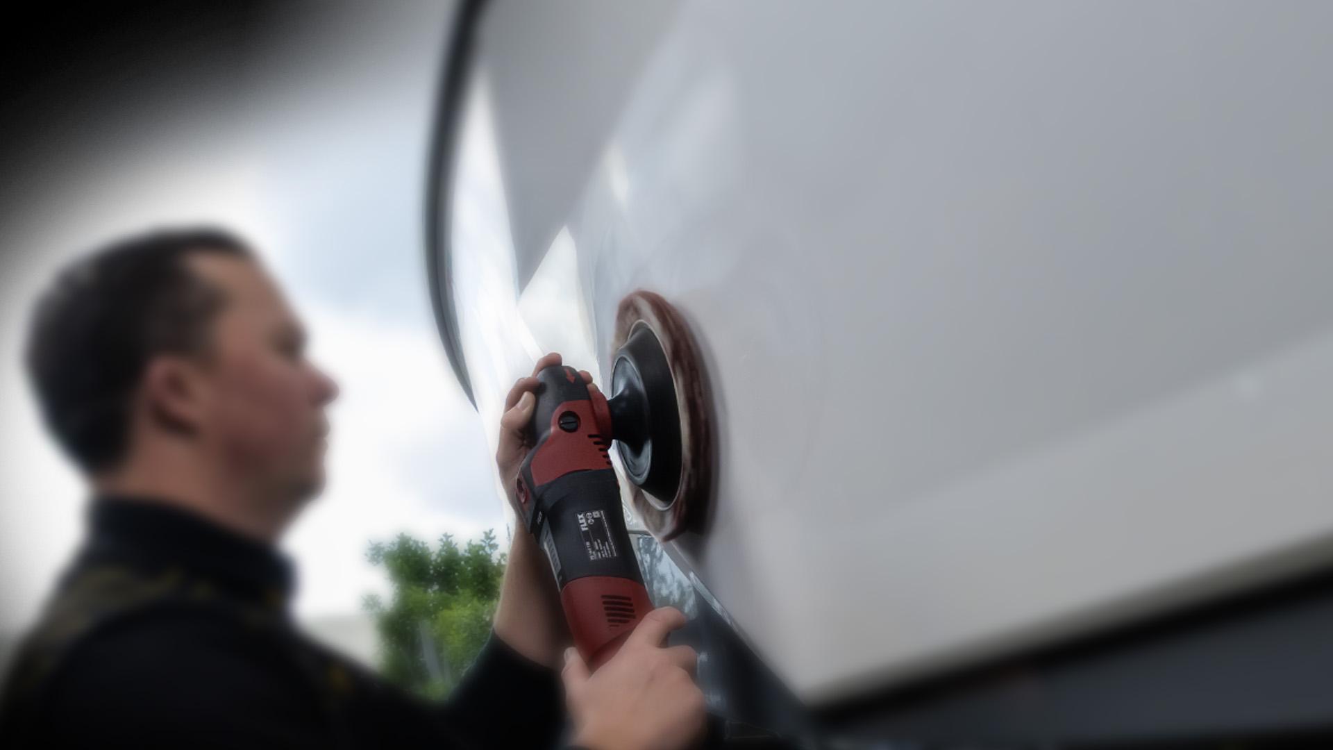 Photograph of a man using a power buffer to polish the side of a white boat. The scene highlights boat maintenance work, with the man focused on smoothing the surface under natural daylight.