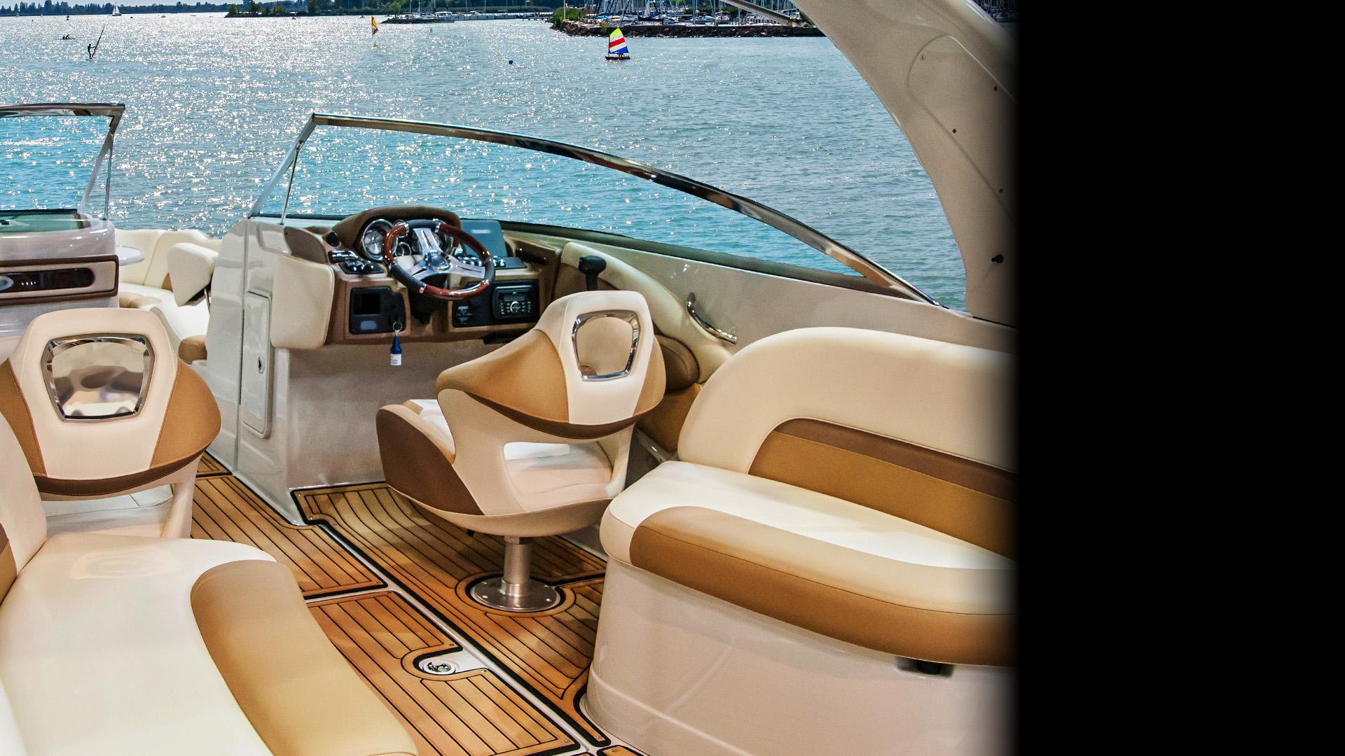 Photograph of a boat's cockpit area showing a beige and brown captain's chair, a steering wheel, and control panel with water visible through the windshield. The interior features wood-patterned flooring and cushioned seating designed for comfort and navigation.