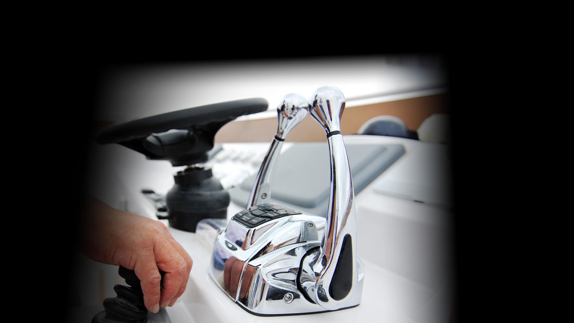 A close-up of a hand operating a boat throttle control with two chrome levers and a steering wheel in the background.
