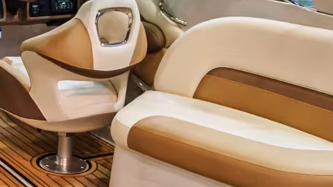 Photograph of a boat's cockpit area showing a beige and brown captain's chair, a steering wheel, and control panel with water visible through the windshield. The interior features wood-patterned flooring and cushioned seating designed for comfort and navigation.