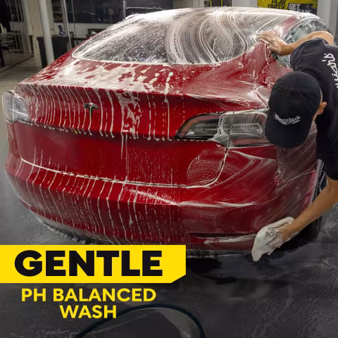 A red Tesla covered in thick soap suds is being hand washed in a detailing garage. Large text reads “GENTLE” and “PH BALANCED WASH.” Meguiar’s branding is visible on the apparel. The image highlights gentle, pH-balanced vehicle cleaning.